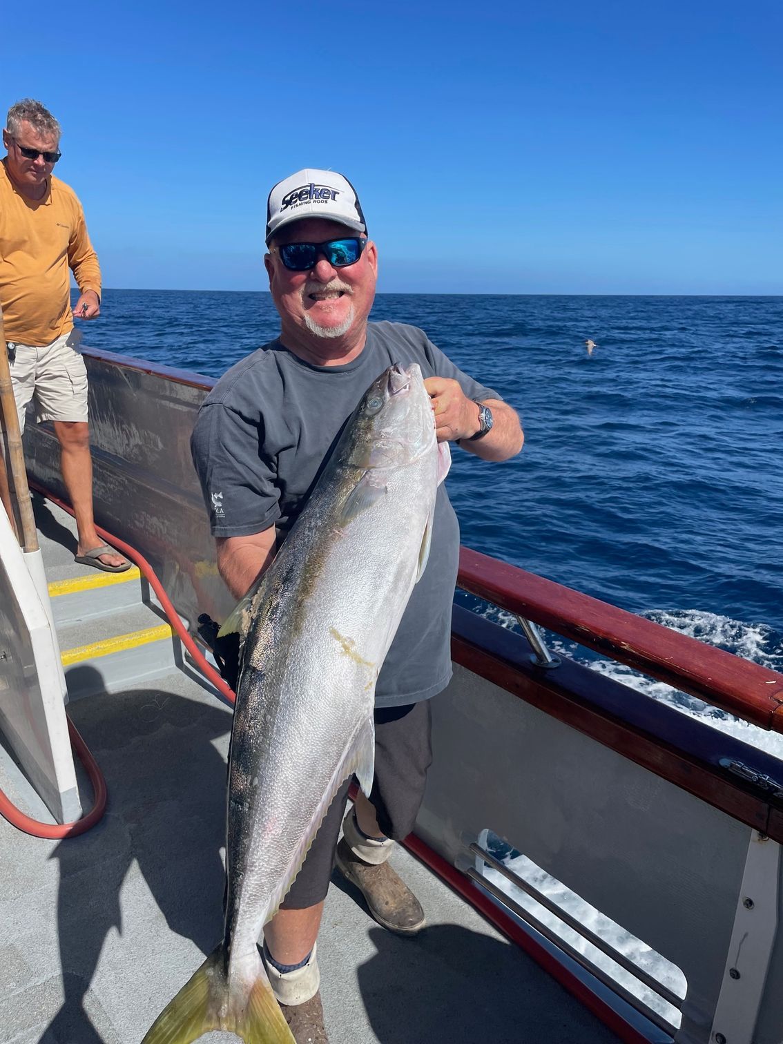 Man on boat holding a large yellowtail fish. Smiling, with a man in the background. Blue water and sky.