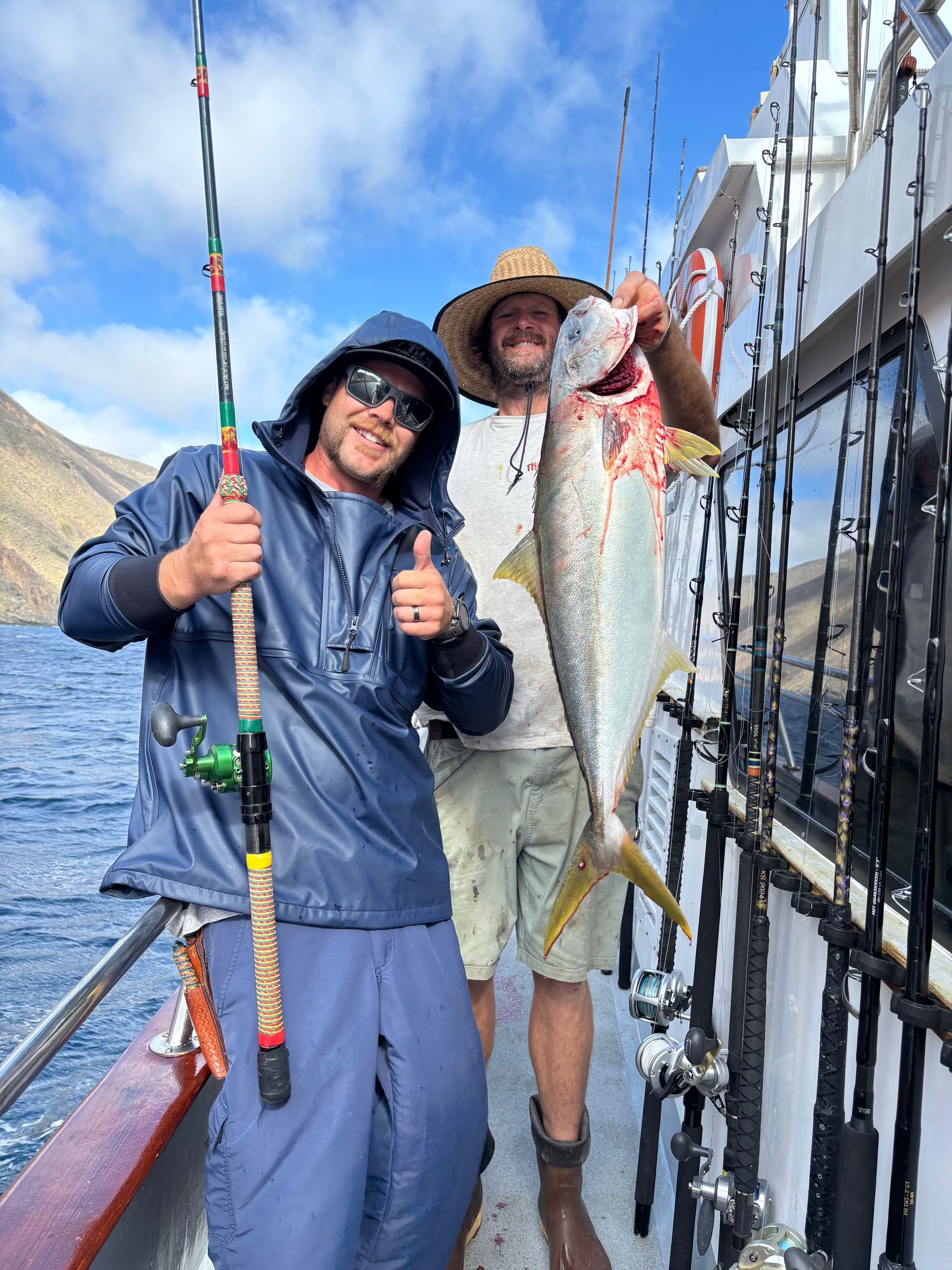Two men on a boat, one holding a large fish, smiling, ocean backdrop.