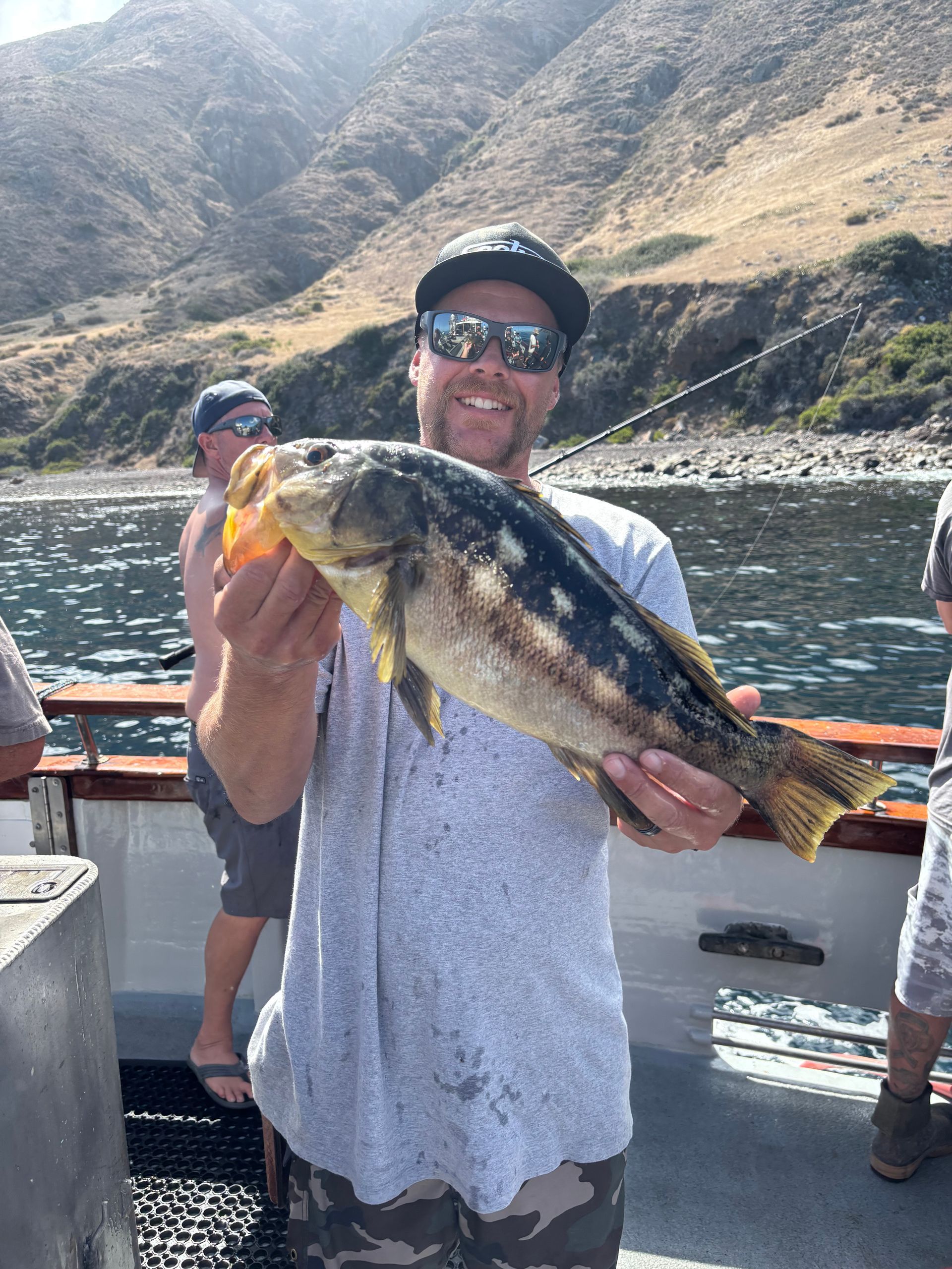 Man on a boat smiling, holding up a large, spotted fish with mountains and water in the background.