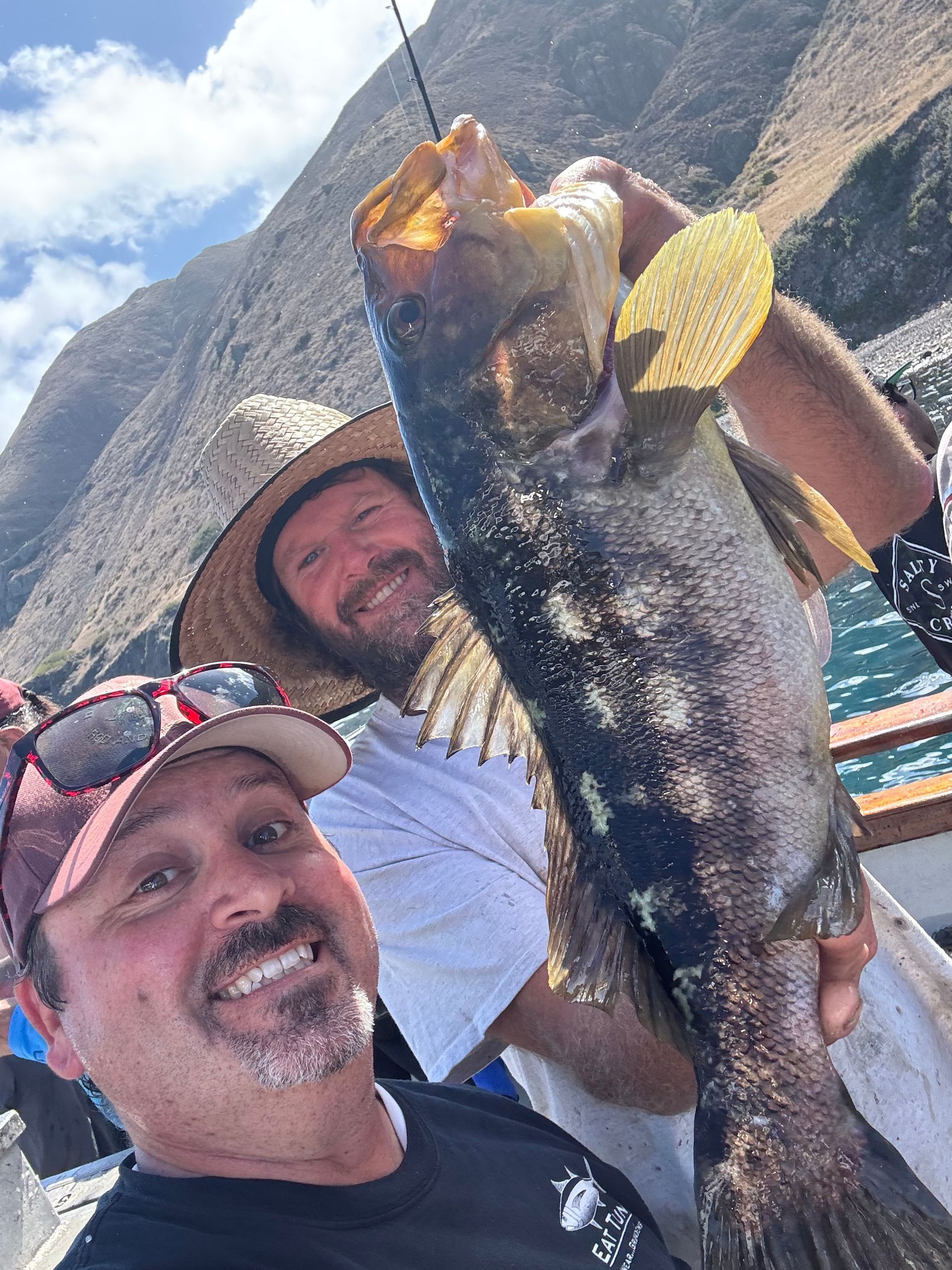 Two men smiling, holding up a large fish on a boat, mountains in the background.