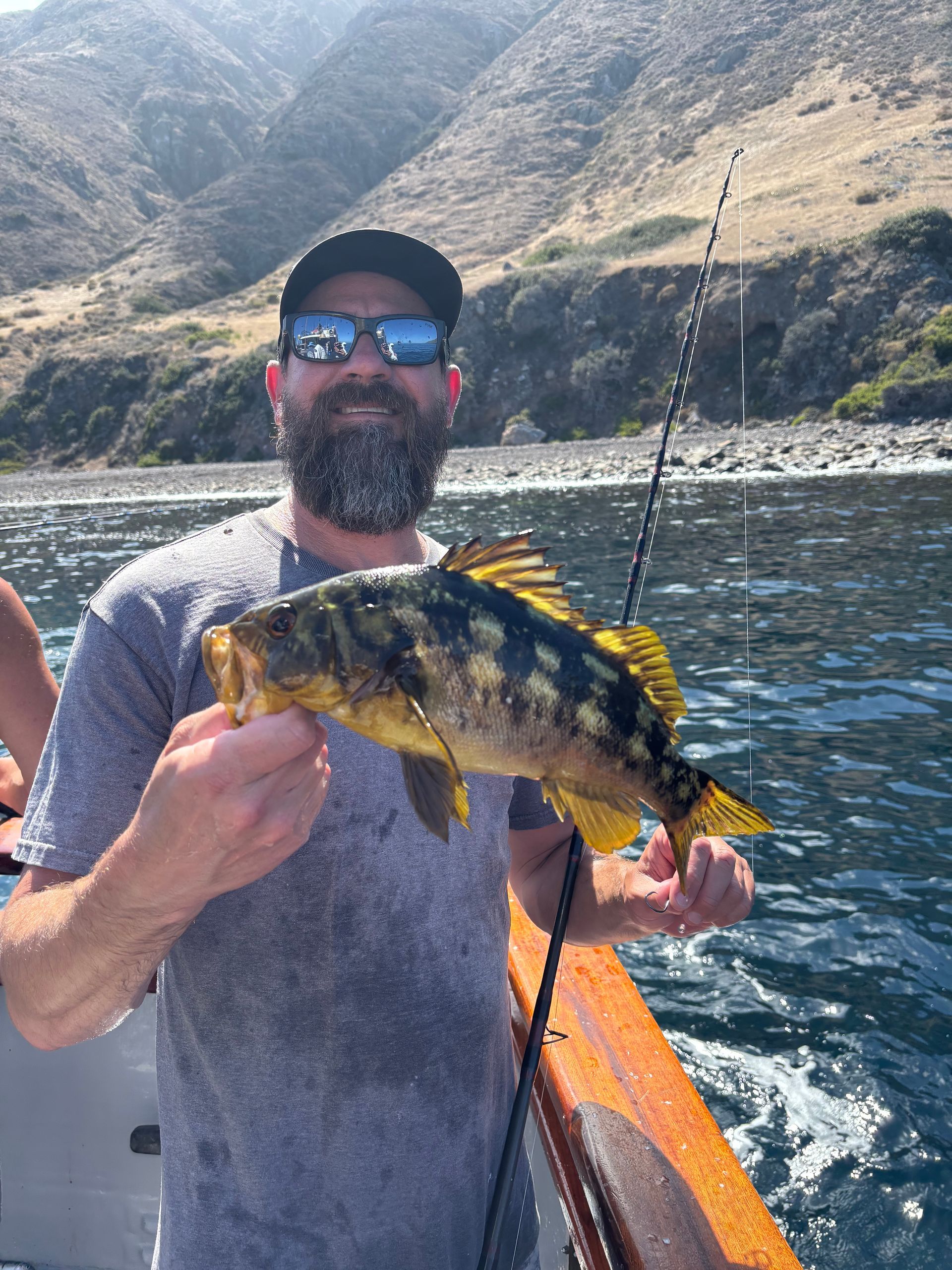 Man on boat, smiles holding a yellow, brown fish. Mountains and sea in background.