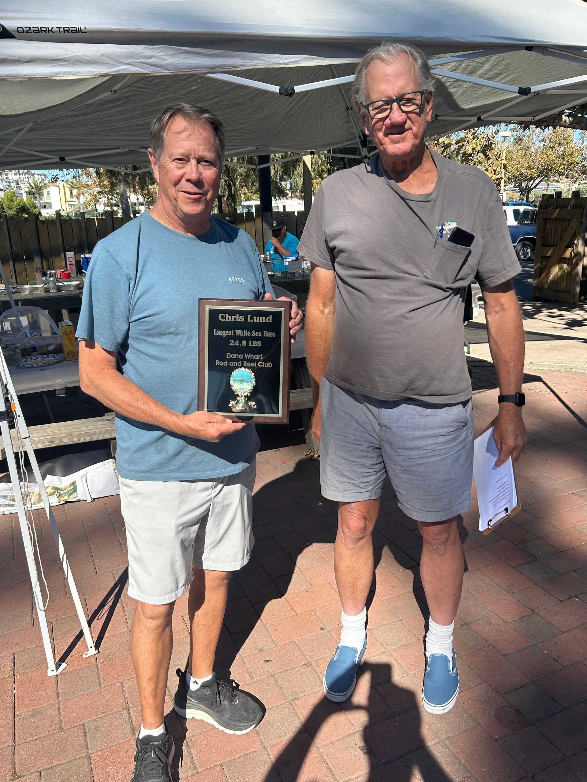 Two men holding a plaque outdoors. One man wears blue and holds the plaque, the other wears gray and shorts.