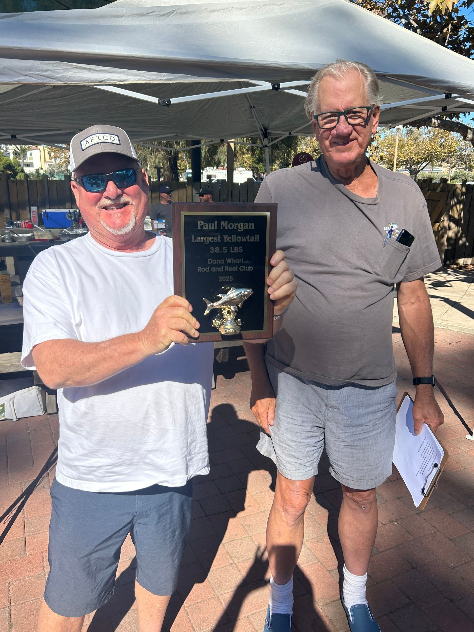 Two men holding an award plaque outside. One in white shirt and sunglasses. Other in gray shirt and shorts.