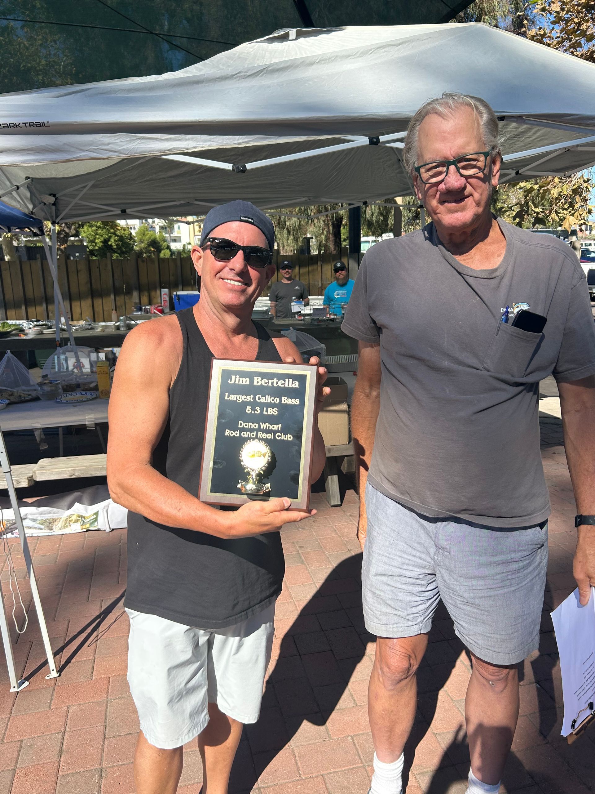 Two men outdoors, one holding a plaque that reads 