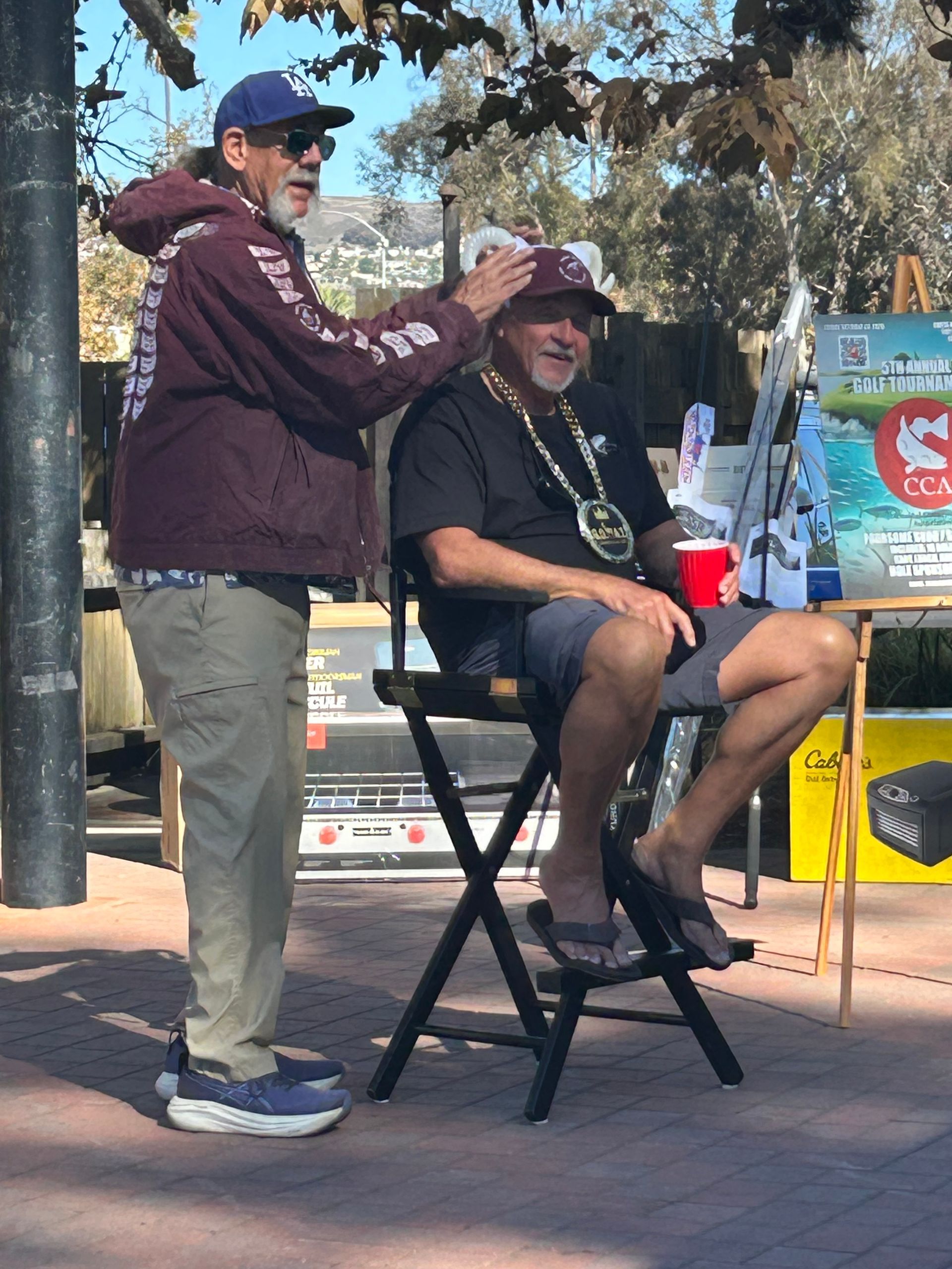 Two men, one in a hoodie touching the other's head, who is seated and wearing a hat, holding a cup. Outdoors.