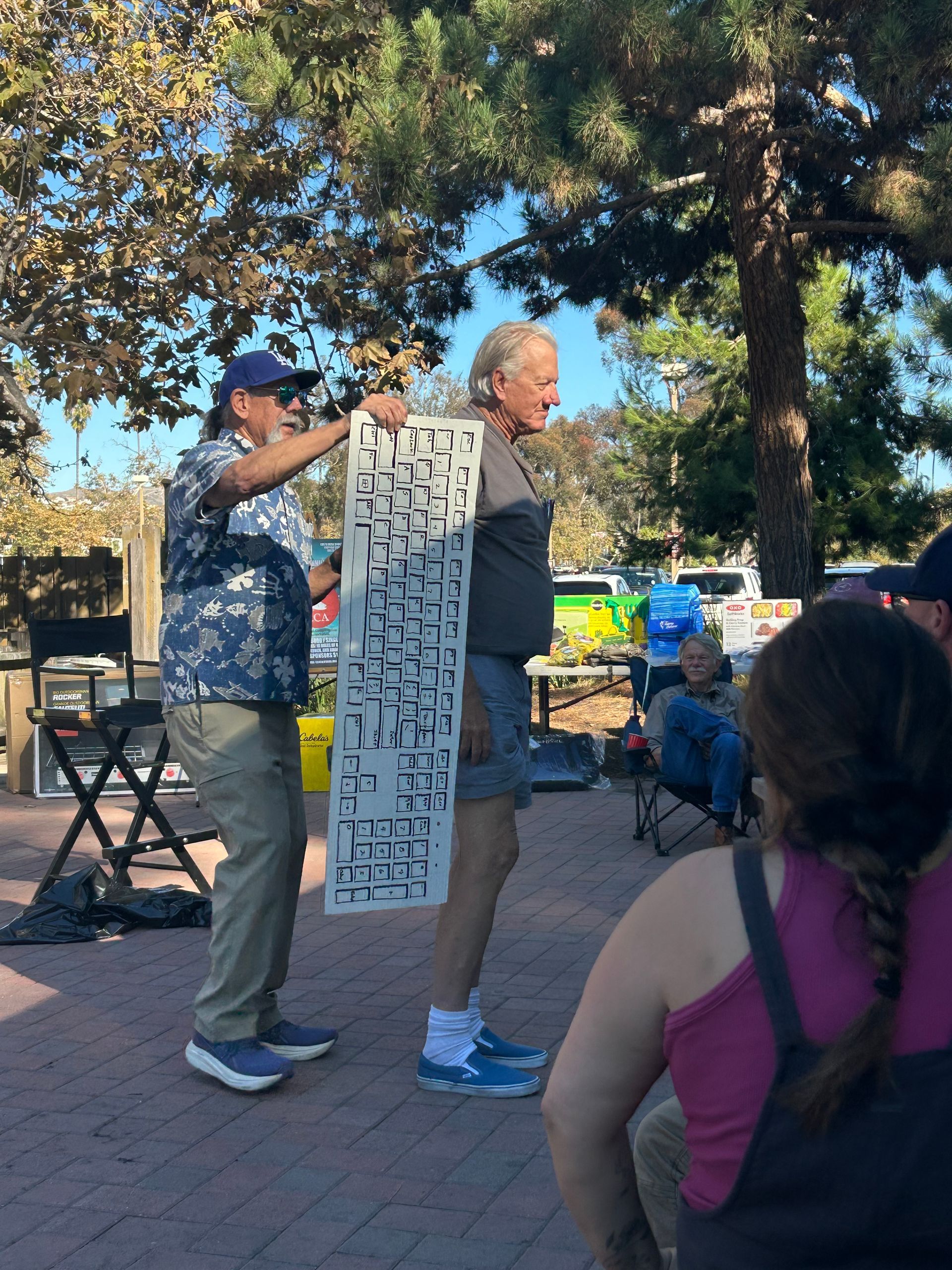 Man holding a stencil against another man standing outdoors. Other people and trees in the background.