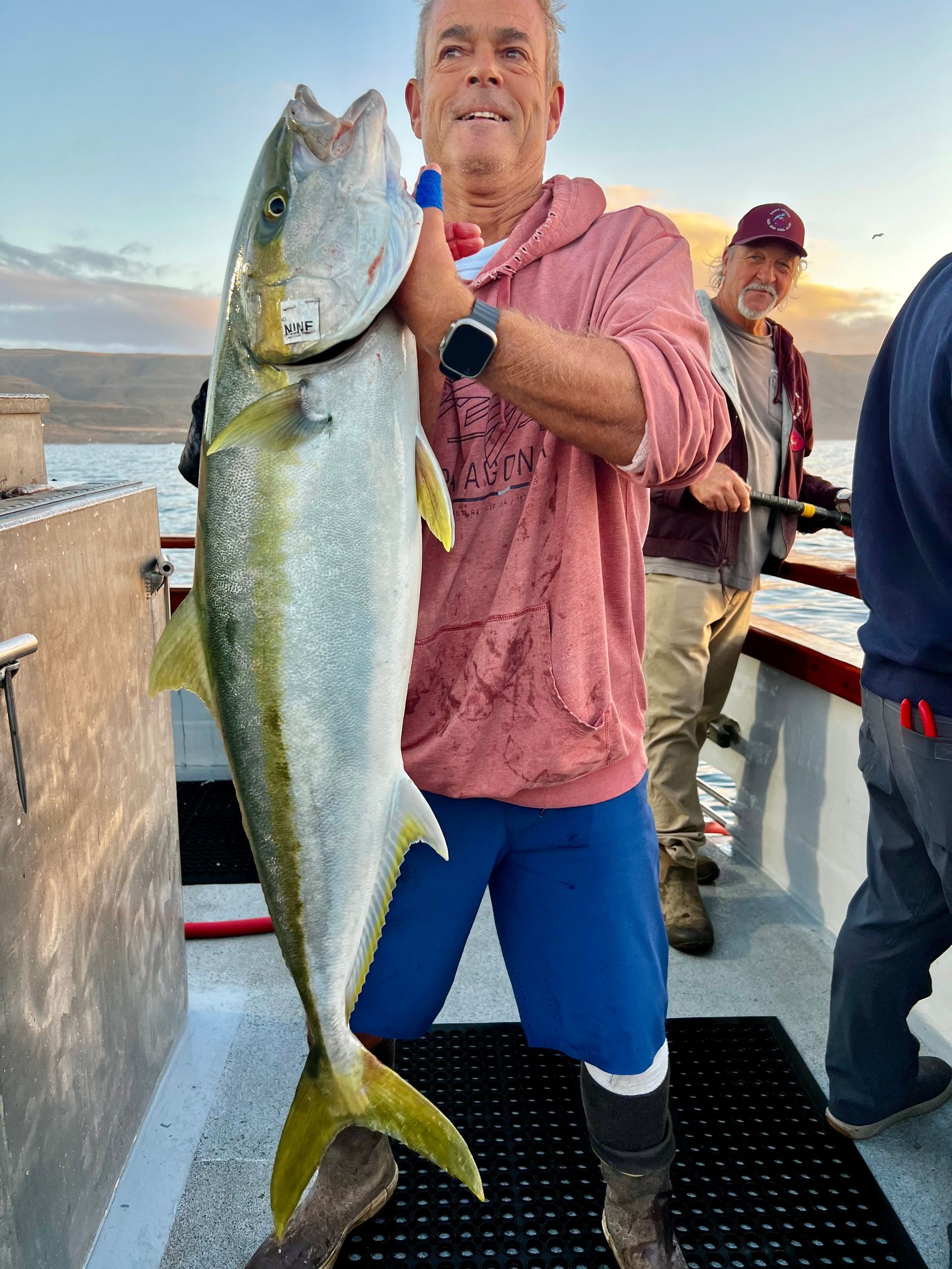 Man holding a large yellowtail fish on a boat, wearing pink hoodie, blue shorts, and smiling. Ocean in the background.