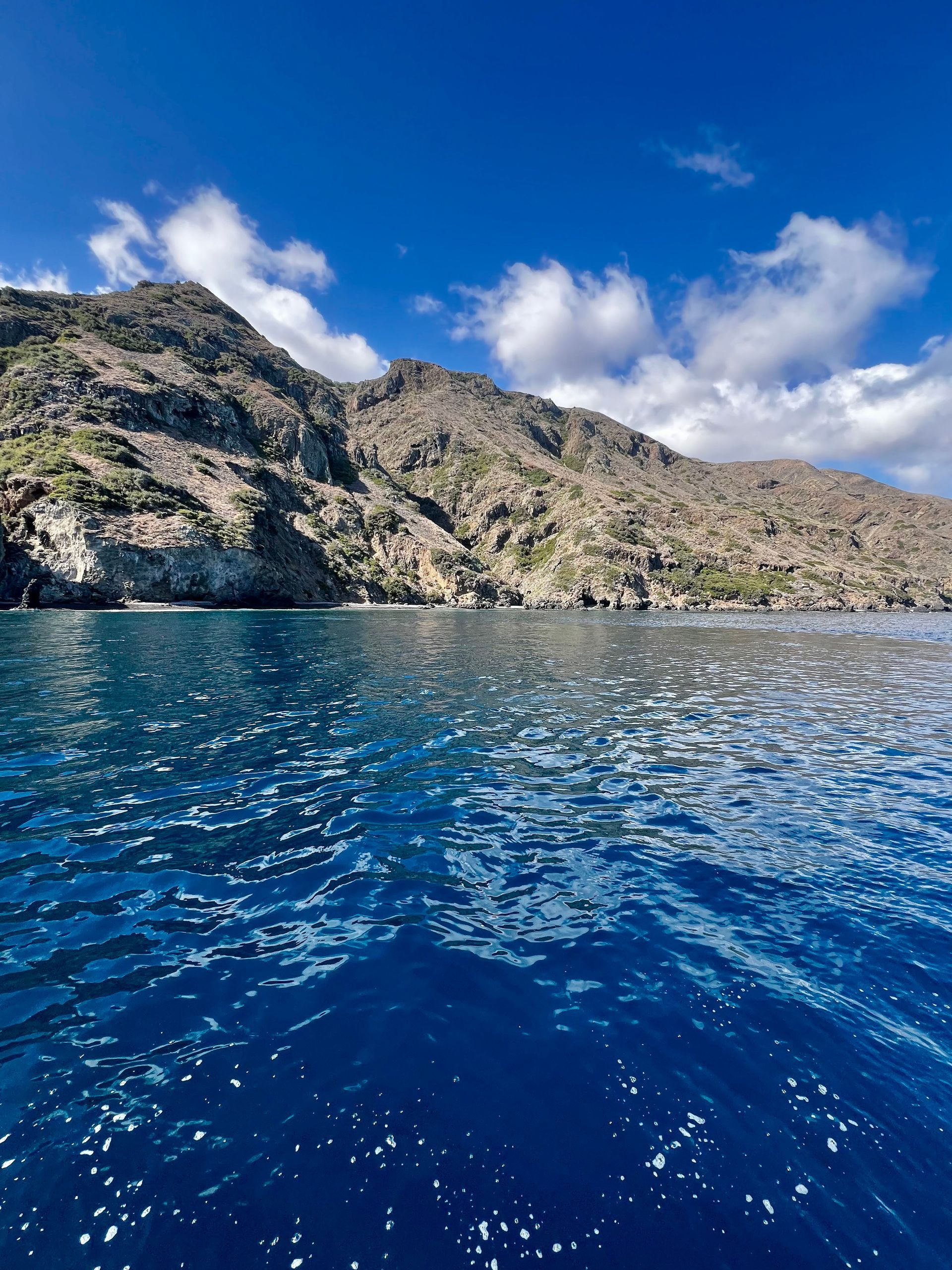 Blue water in foreground, rocky mountain under a bright blue sky with fluffy clouds.
