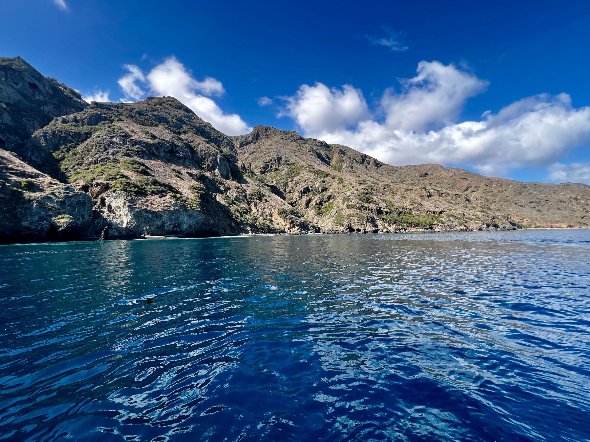 Blue ocean water with a mountainous coastline under a bright blue sky with clouds.