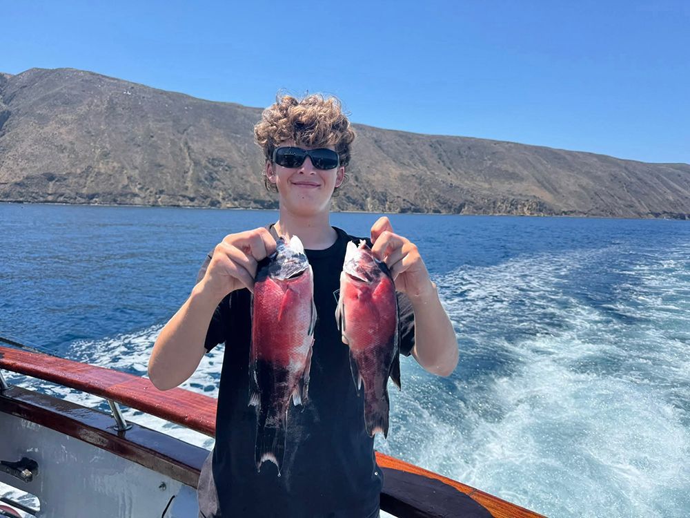 Young person on a boat holding up two caught pink fish, smiling, with mountains and water in the background.