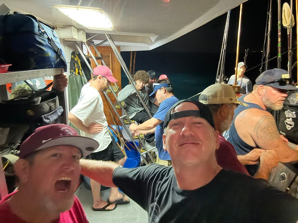 Men on a fishing boat at night. Some are holding rods, others smiling.