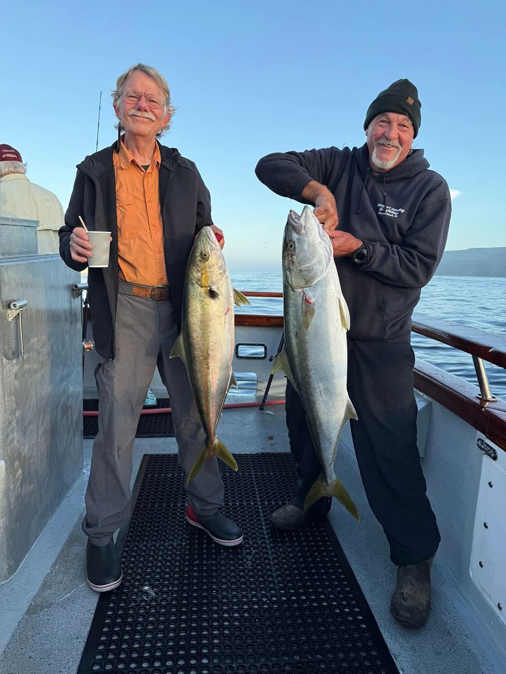 Two men on a boat holding up large yellowtail fish. One man smiling, the other holding a coffee.