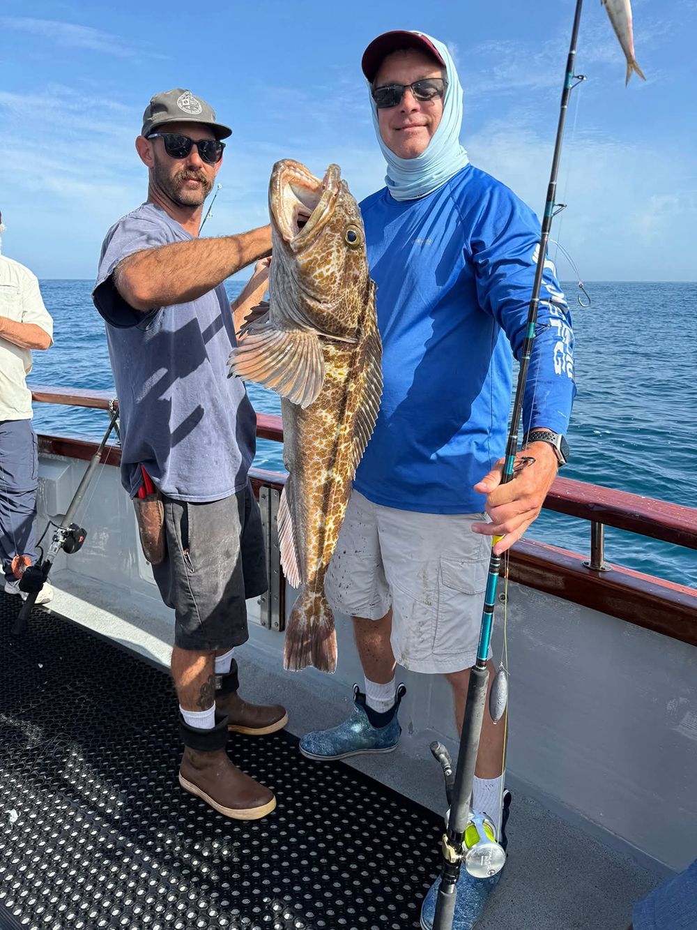 Two men on a boat holding a large brown fish. Sunny day, blue water.