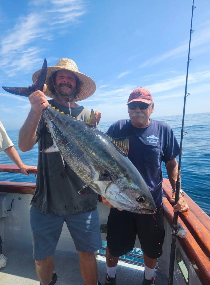 Two men on a boat holding a large tuna fish they caught. Blue sky and water visible.