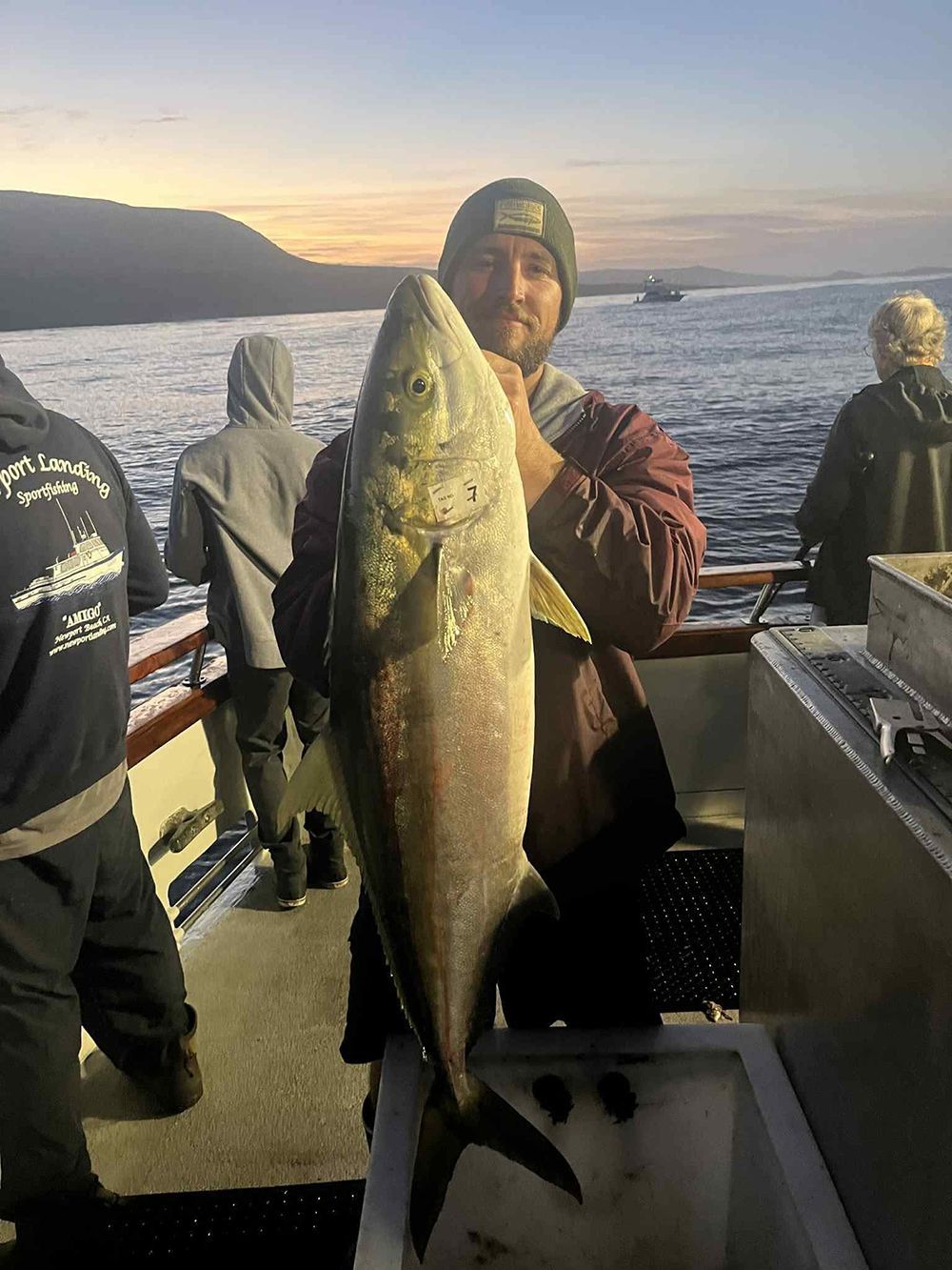 Man on a boat holding up a large fish. Sunset on the water, other people fishing.