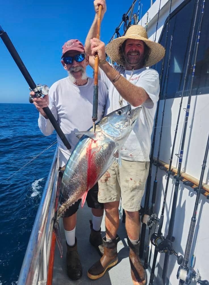 Two men on a boat hold up a large fish. Sunny day; one man wears a straw hat, the other a cap.