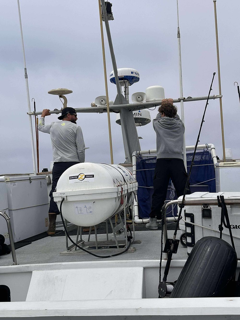 Two people on a boat adjusting equipment near a radar tower under a cloudy sky.
