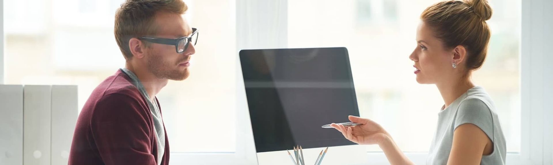 A person in a red sweater looks at a coworker in a grey top, who points toward a computer monitor in a bright office.