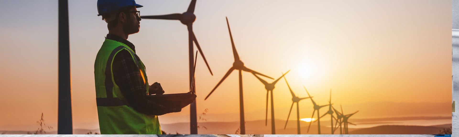 Worker in safety gear beside wind turbines at sunset, with a warm orange sky.