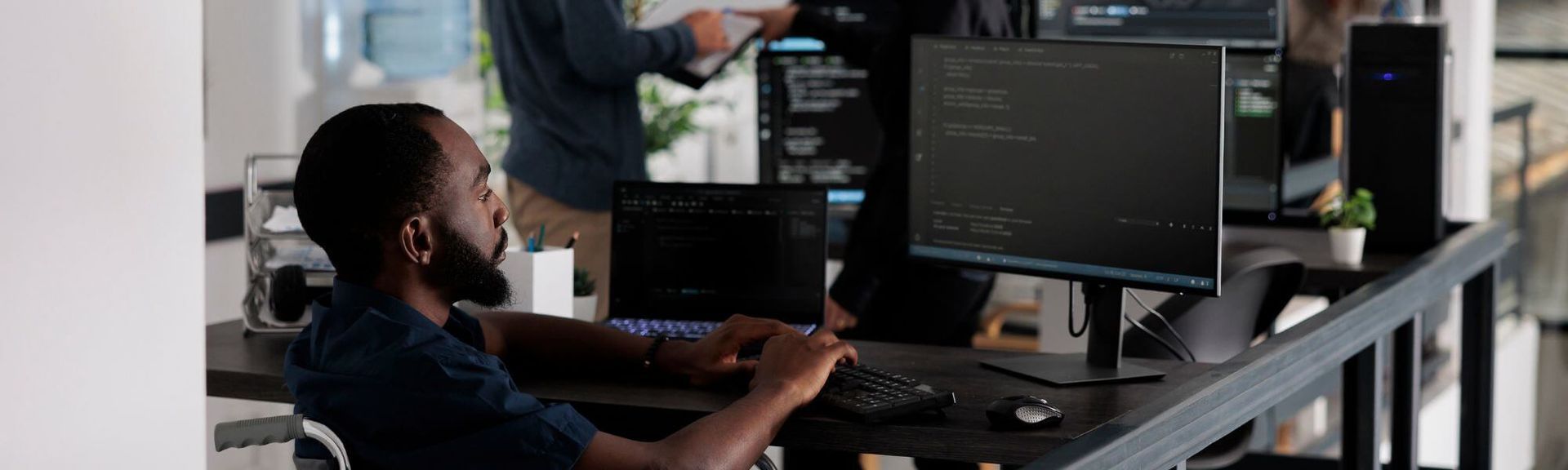 A programmer in a wheelchair works at a desk with multiple monitors displaying code in a modern office.