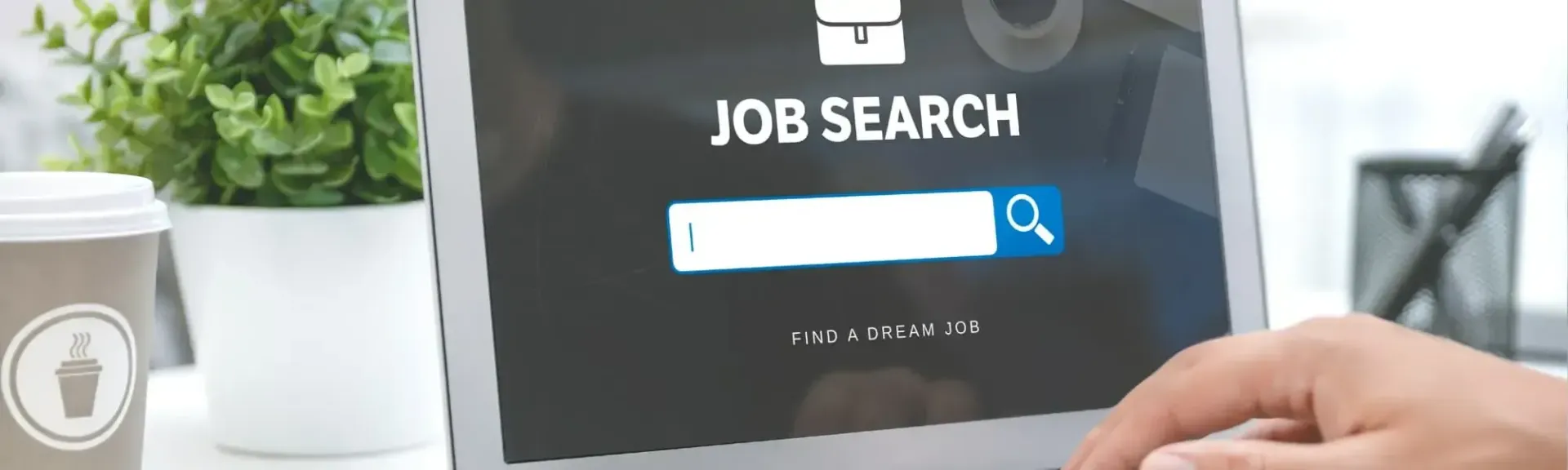 A person searching for jobs on a laptop screen with a coffee cup and a small potted plant on a desk.