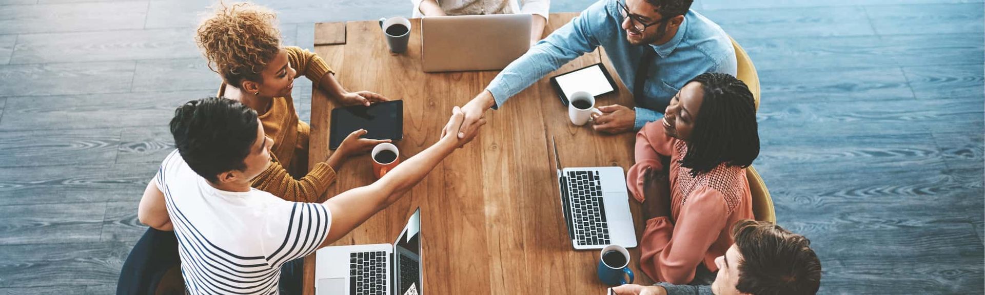 Team meeting around a wooden table with laptops, taking a celebratory group fist bump outdoors near water