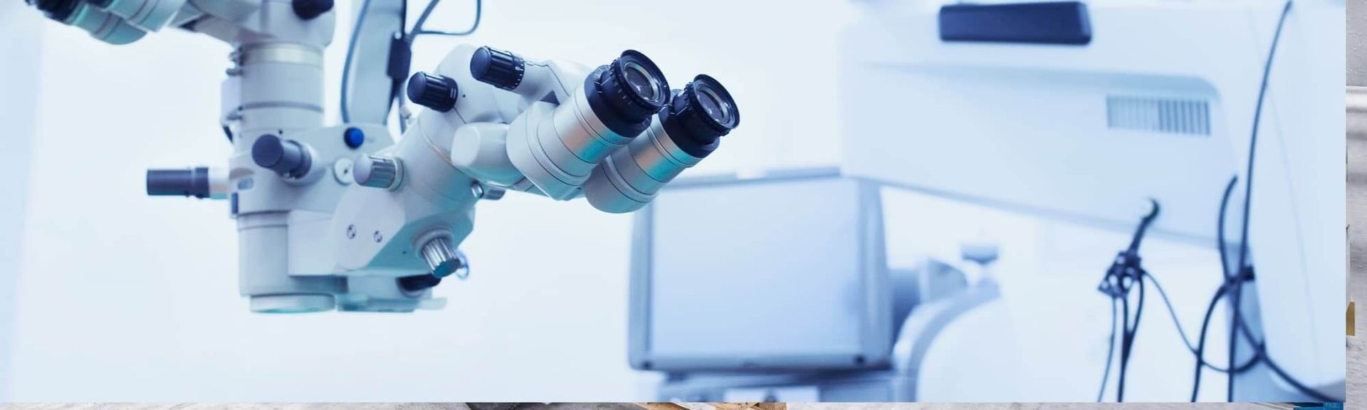 A close-up of a medical operating microscope with dual eyepieces in a bright, clinical environment.
