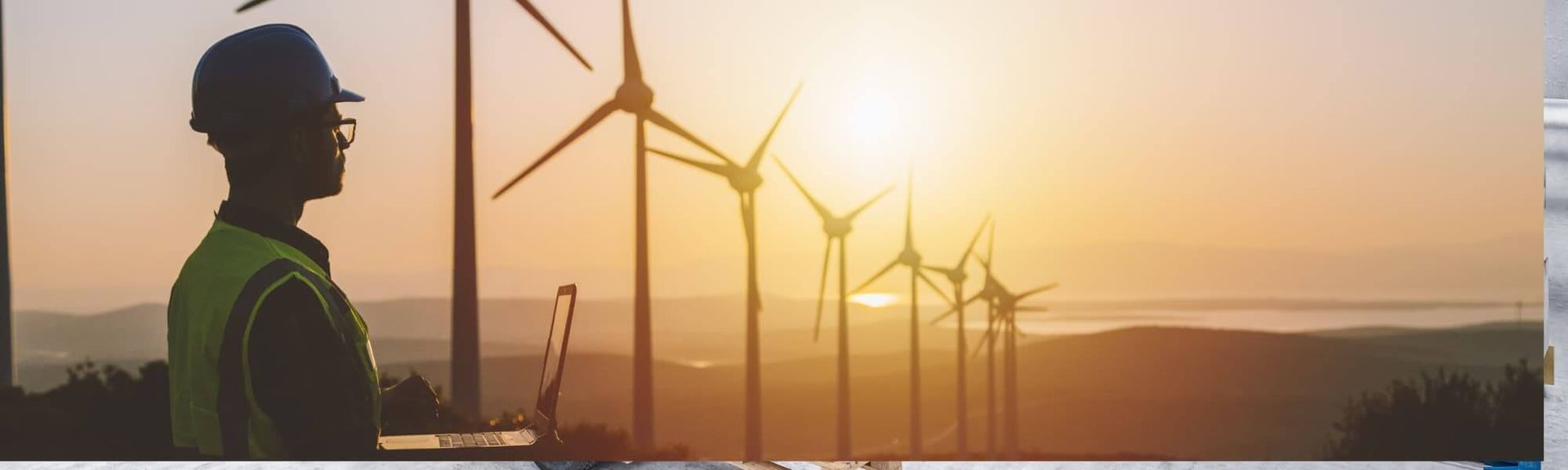 A silhouette of a worker in a safety vest and hard hat overlooking a field of wind turbines at sunrise.