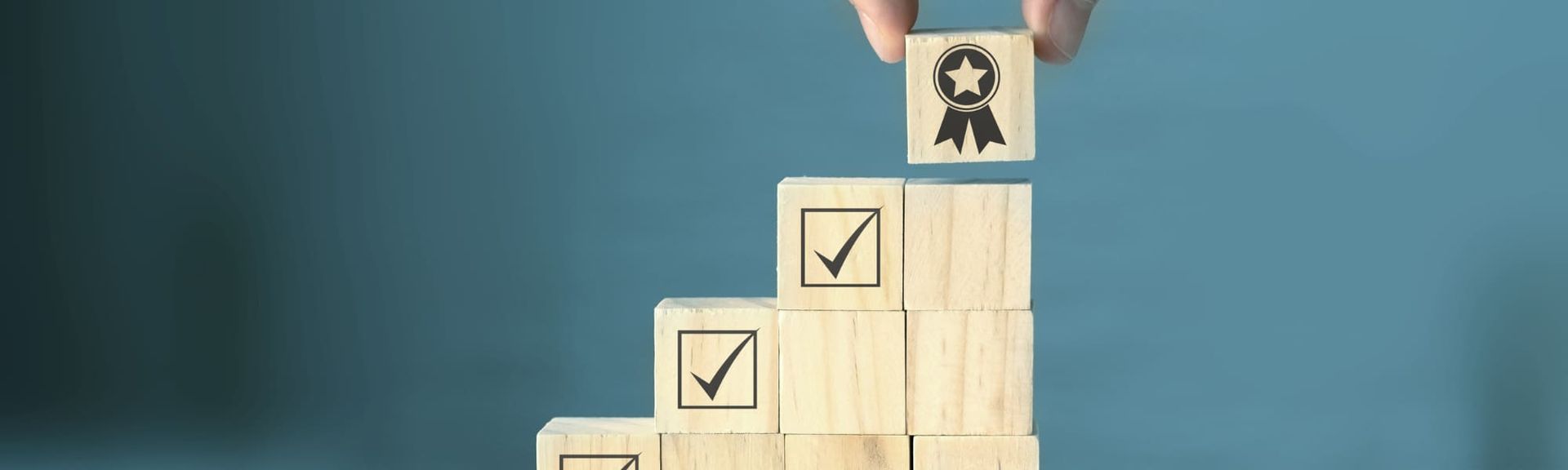 A hand places a wooden block with a trophy icon atop a stepped stack of blocks marked with checkmarks.