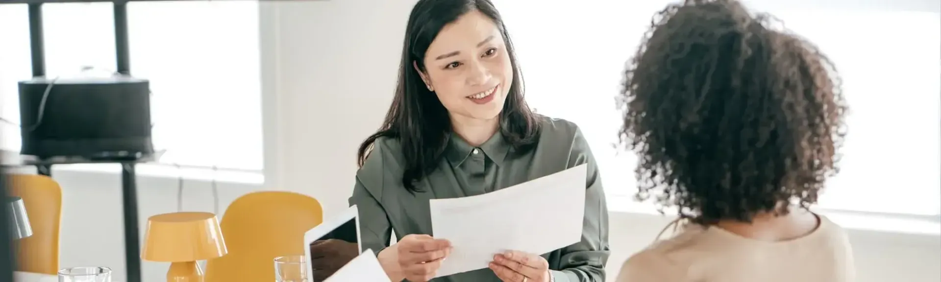 Two professionals sitting at a table in a bright office, with one person handing a document to the other.