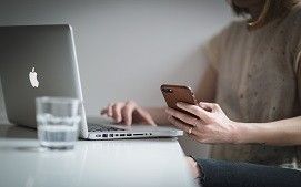 A person sits at a desk with an open laptop, holding a smartphone in their hand with a glass of water nearby.