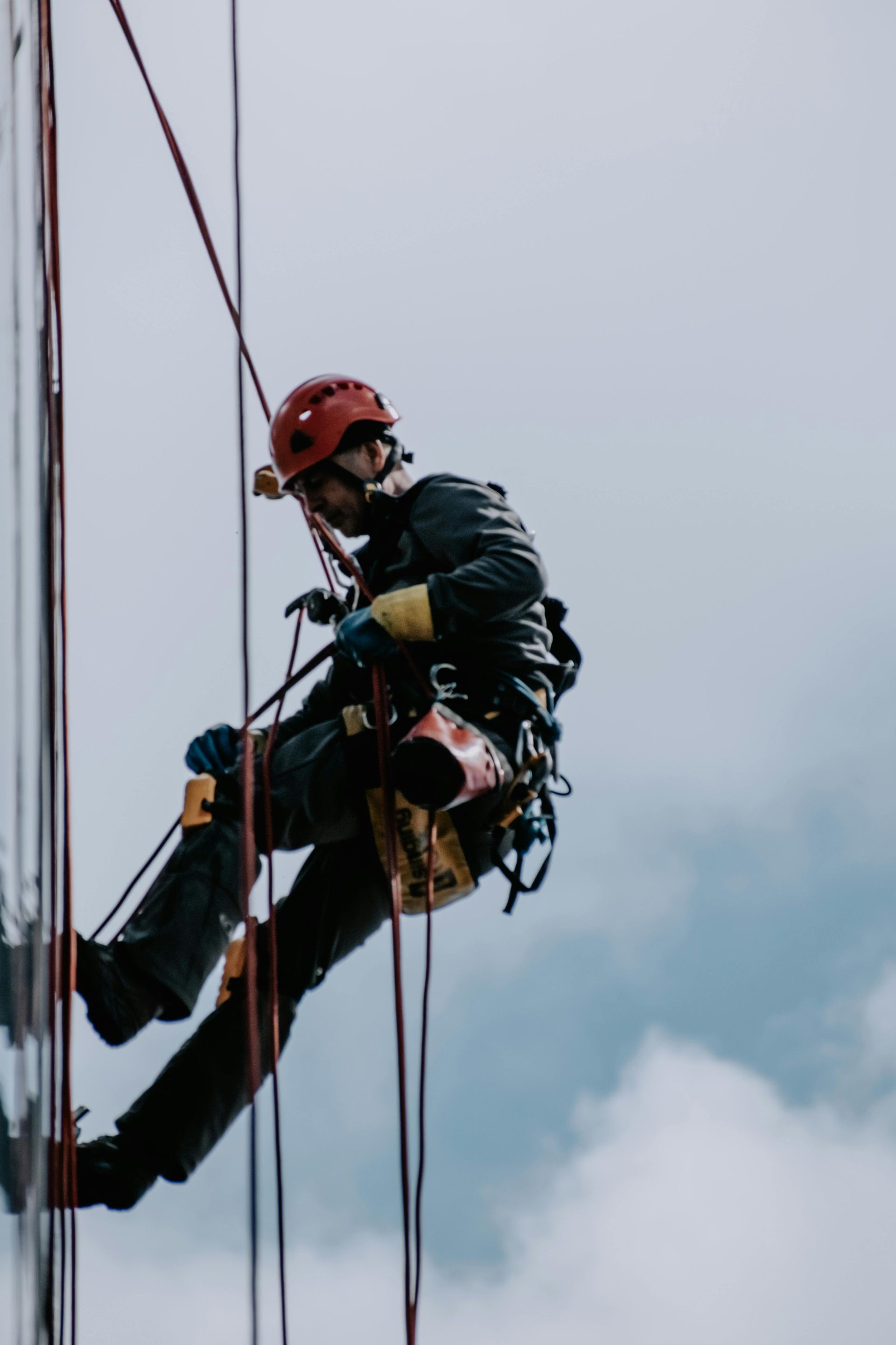 A worker in a red helmet and climbing gear rappels down the side of a building against a cloudy sky.
