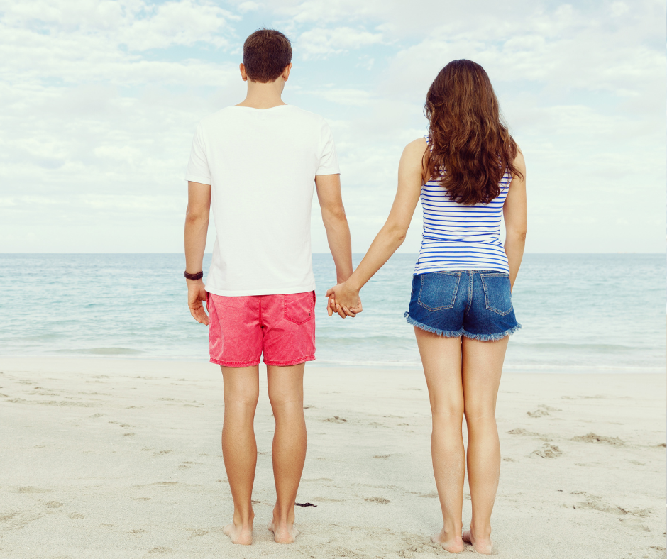 A man and a woman are holding hands on the beach and looking at the ocean.