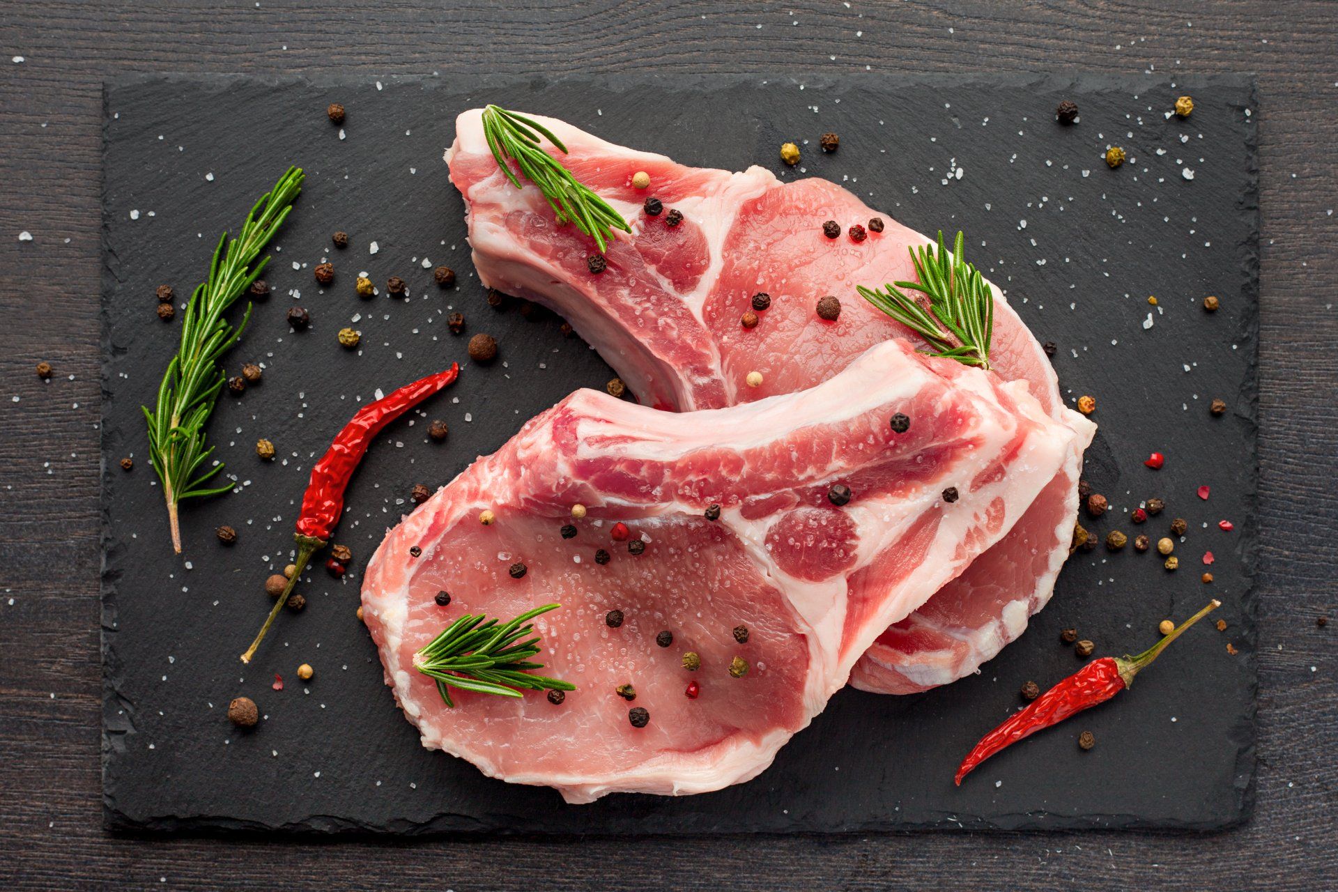 Two raw pork chops on a black cutting board with peppers and herbs.