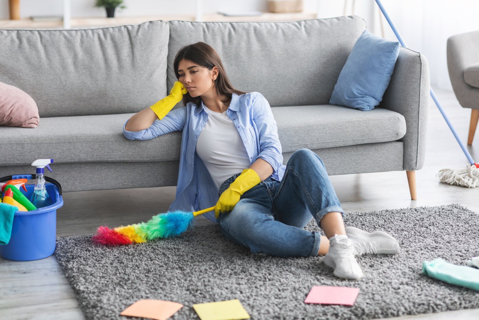A woman is sitting on the floor in front of a couch holding a duster.
