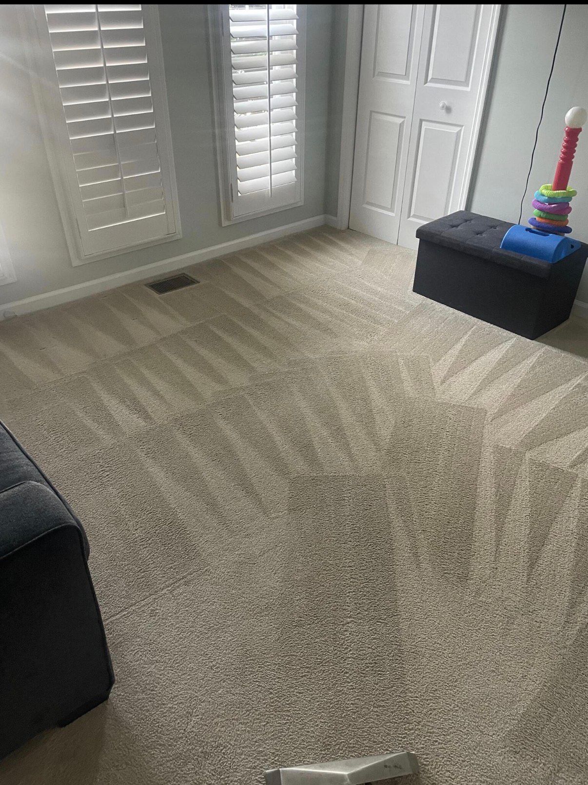 Beige carpet in a room with a window, white shutters, and a black storage ottoman with toys on top.
