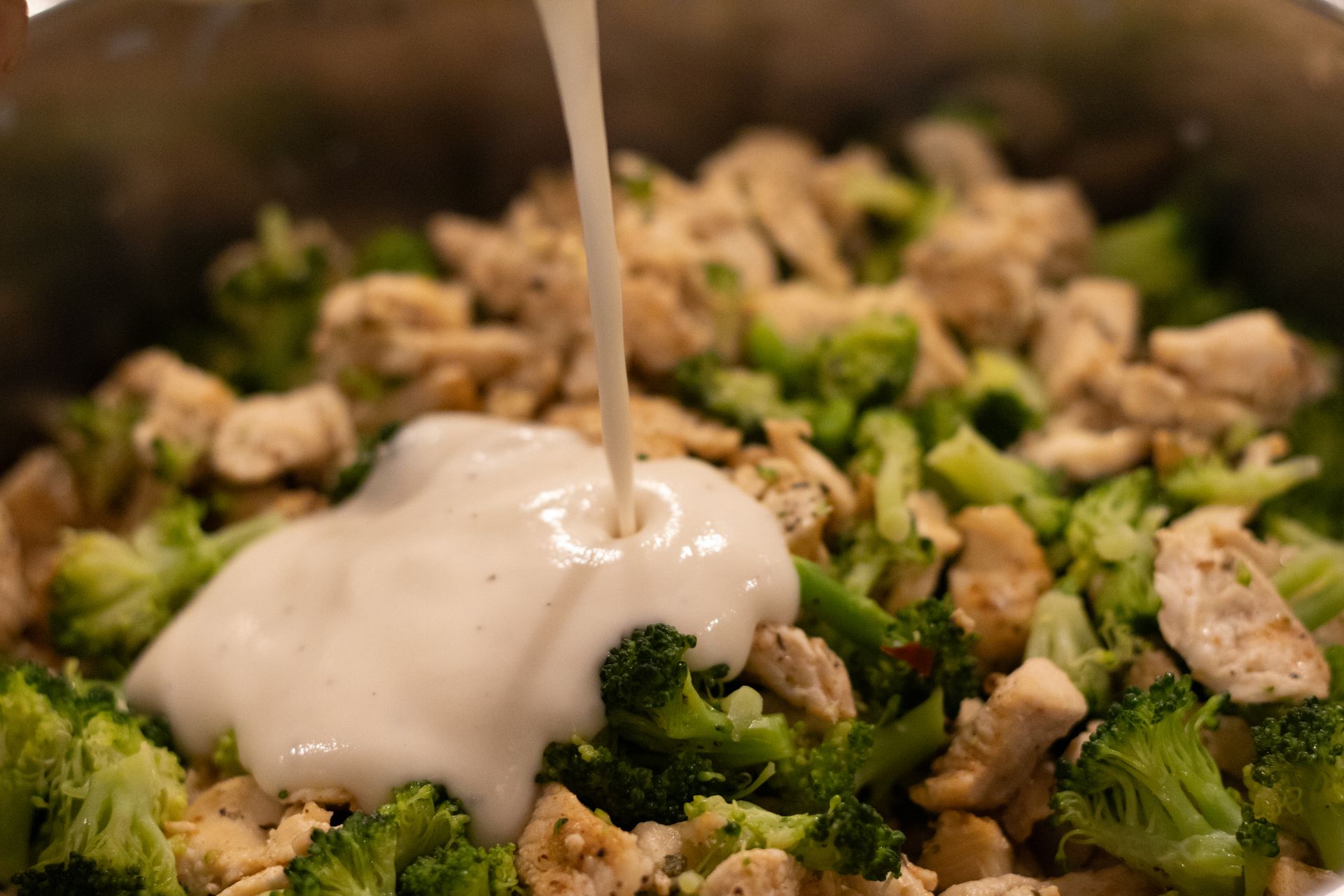 Alfredo sauce being poured into a pot filled with pasta, chicken, broccoli