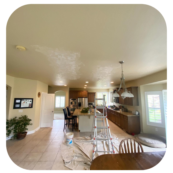 A kitchen undergoing ceiling painting, featuring a ladder, drop cloths, and patches of light paint on a tan ceiling.