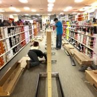 Two people working on a floor-level display structure in the aisle of a retail store filled with shoe boxes on shelves.