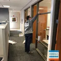A person in a gray hoodie cleans a glass office door in a professional workspace.