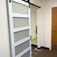 A white, five-panel frosted glass sliding barn door installed on a black track in a room with a closed wooden door.