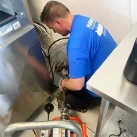 A person in a blue t-shirt kneeling on the floor to work on plumbing equipment behind a stainless-steel appliance.