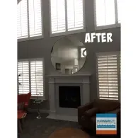 A living room featuring a fireplace with a large round mirror above it, flanked by windows with white plantation shutters.