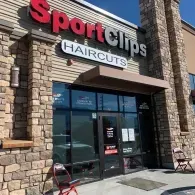 Exterior view of a Sport Clips Haircuts storefront with stone siding, glass doors, and a red and white sign.