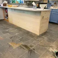 A kitchen island with a light-wood panel exterior stands on a grey tiled floor with visible patches of wet mortar.