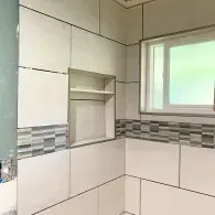 A tiled shower wall with large cream tiles, a horizontal mosaic accent strip, a built-in recessed shelf, and a window.