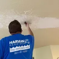 A handyman in a blue shirt scrapes a popcorn texture off a ceiling with a metal tool.