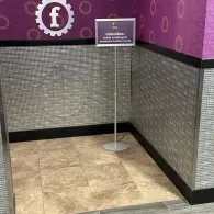 An informational sign on a metal stand in front of a purple wall with a logo, set against silver textured wainscoting.