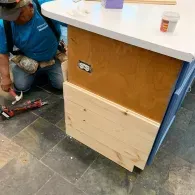 A person kneeling on a tiled floor installs wood paneling onto the side of a kitchen island with an electrical outlet.