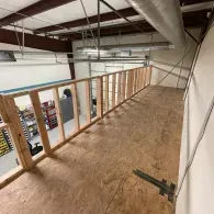 A wooden wall frame under construction serves as a railing along the edge of an elevated loft in a warehouse setting.